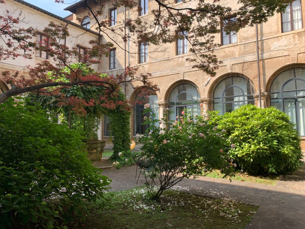 L'immagine rappresenta una vista in diagonale del chiostro della Biblioteca Bertoliana a Vicenza. Sono presenti alberi e fiori di vario tipo, illuminati parzialmente dal sole che entra all'interno dello spazio del chiostro.